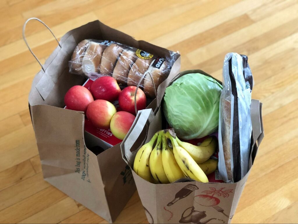 Duas sacolas de papel no chão de madeira com compras de mercado contendo frutas, banana, maçã, verduras e pão