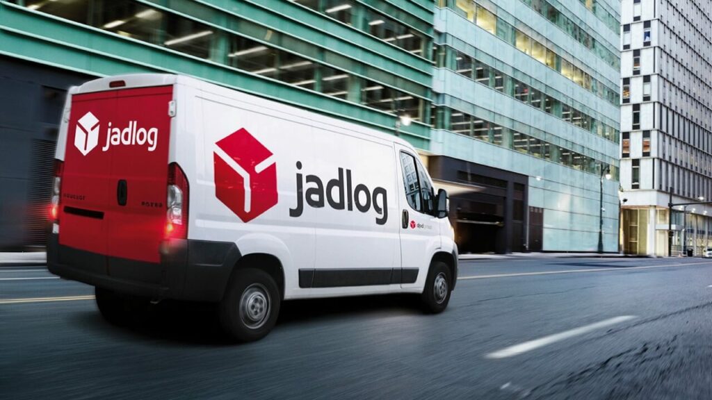 Veículo branco e vermelho da transportadora Jadlog em movimento em uma rua repleta de prédios
