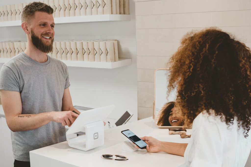 Homem branco com camisa cinza, sorrindo e fazendo o pagamento de um produto, ao lado de uma mulher negra com camisa branca que está sorrindo com o celular na mão pois sabe como fidelizar clientes.